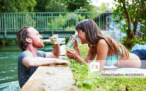 Junger Mann im Fluss  junge Frau liegt im Gras am Flussufer  Mann trinkt aus Bierflasche