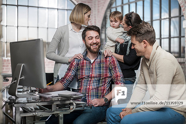 Gruppe von Erwachsenen sitzt um den Computer herum und diskutiert  eine Frau hält ein Kleinkind hinter sich