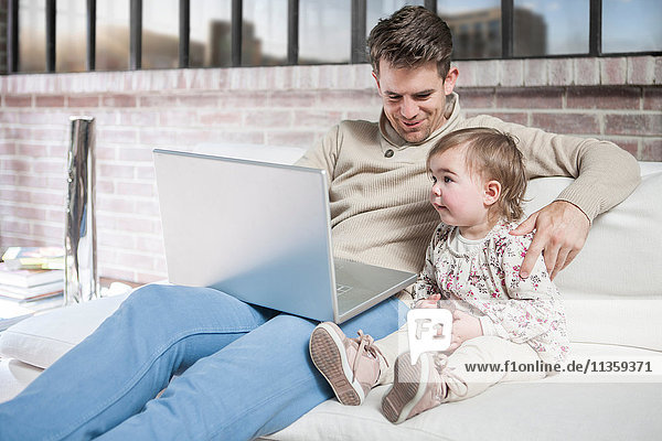 Vater sitzt mit seiner kleinen Tochter auf dem Sofa und schaut auf den Laptop