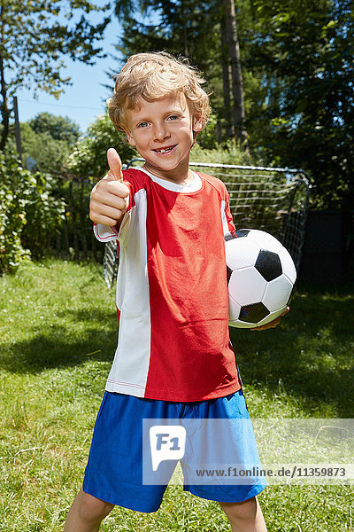 Porträt eines Jungen in Fussballuniform  der im Garten einen Fussball hält und Daumen hochhebt