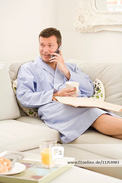Erwachsener Mann  der auf dem Sofa liegt  telefoniert und frühstückt.