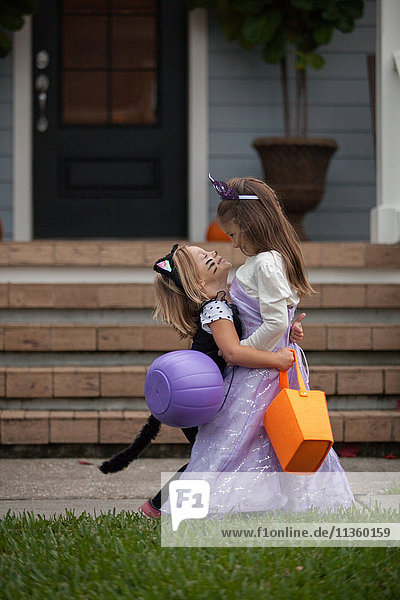 Two sisters trick or treating in cat and fairy costumes hugging at porch stairway