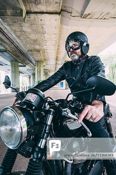 Mature male motorcyclist sitting on motorcycle wearing crash helmet under flyover
