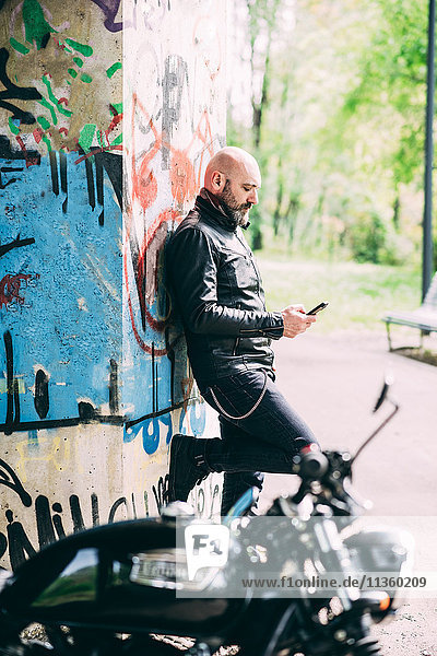 Mature male motorcyclist leaning against wall reading smartphone texts