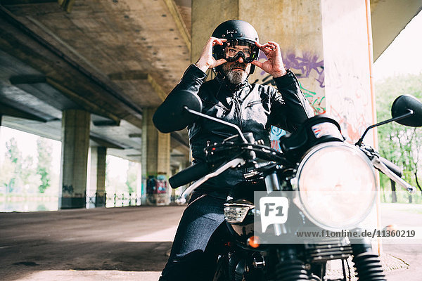 Mature male motorcyclist sitting on motorcycle putting on goggles under flyover