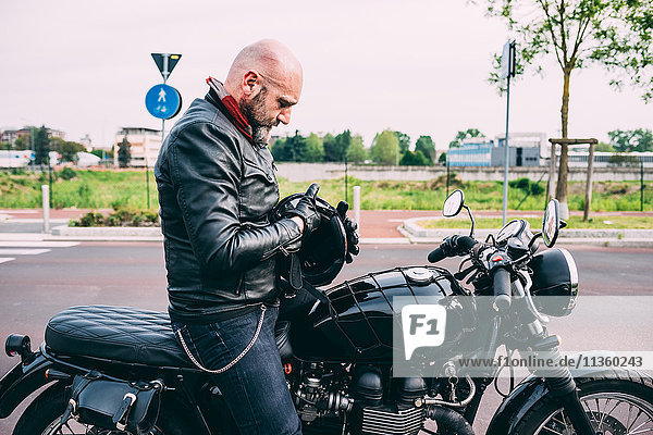 Mature male motorcyclist sitting on motorcycle examining helmet