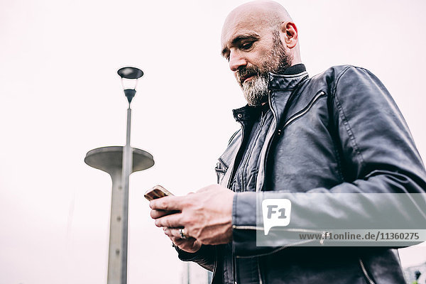 Reifer männlicher Motorradfahrer liest Smartphone-Text vor grauem Himmel