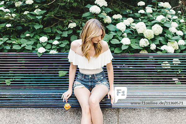 Young woman sitting on bench holding yellow rose looking away