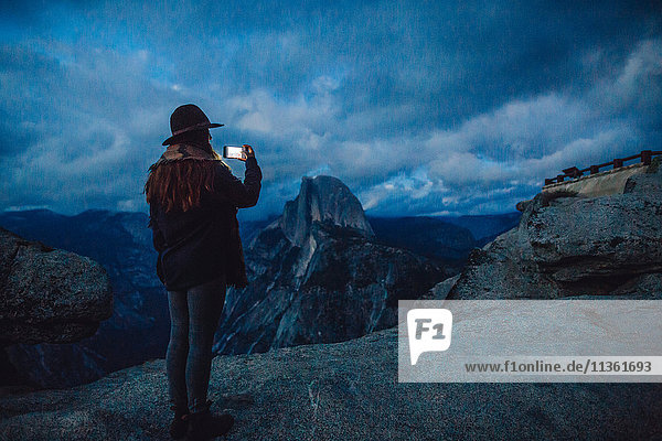 Young woman taking photograph on rock overlooking Yosemite National Park at dusk  California  USA