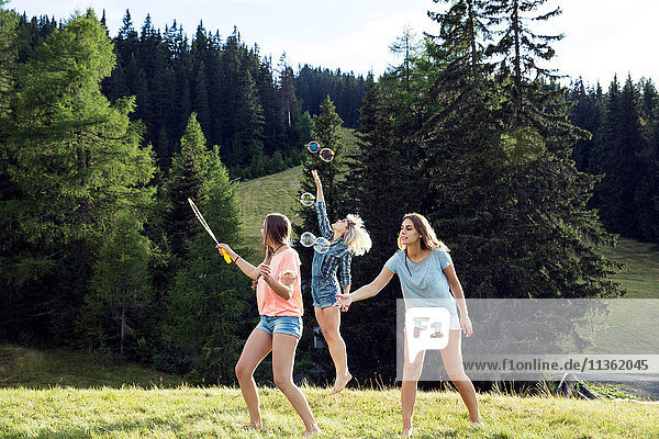 Three female adult friends blowing and jumping for bubbles in field  Sattelbergalm  Tirol  Austria