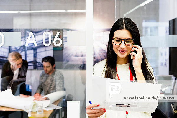 Young woman in office holding paperwork using mobile telephone