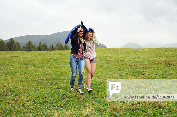 Zwei junge Frauen rennen bei Regen mit einem Anorak in der Hand den Berg hinunter  Sattelbergalm  Tirol  Österreich