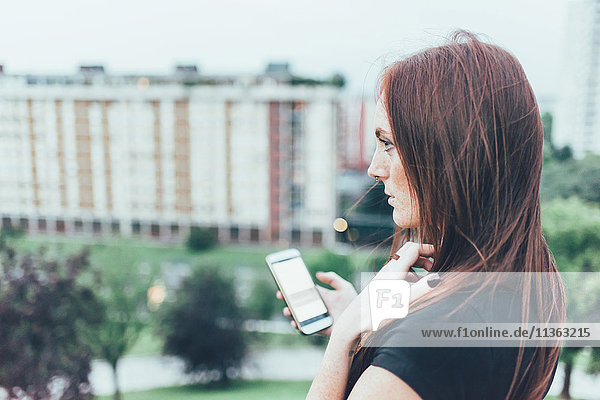 Junge Frau mit langen roten Haaren hält Smartphone und blickt über die Stadt