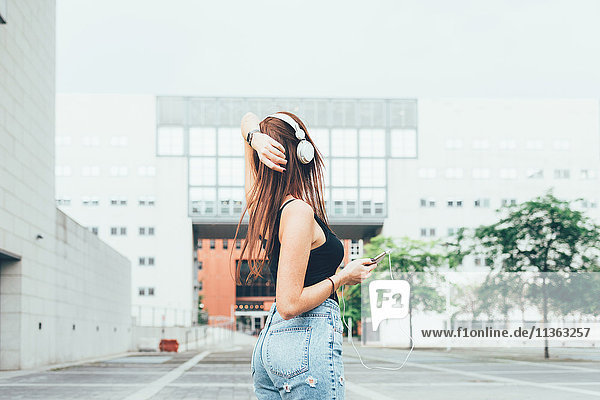 Junge Frau hört Kopfhörer mit der Hand am Kopf vor dem Bürogebäude
