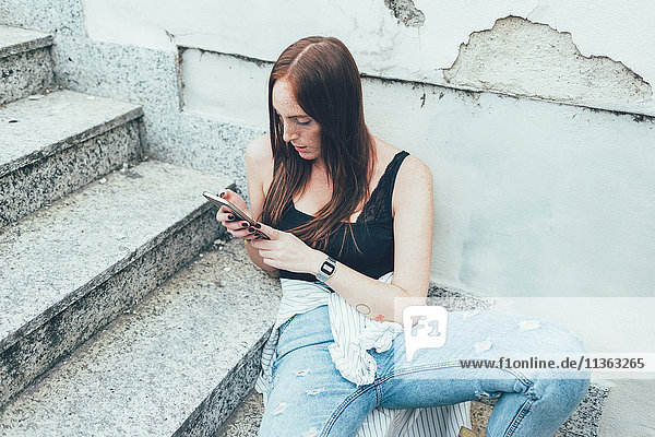 Junge Frau sitzt auf der Treppe und liest Smartphone-Texte.