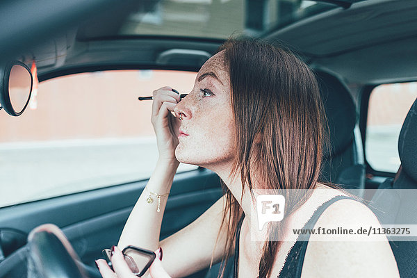 Young woman with freckles applying eyeshadow in car mirror