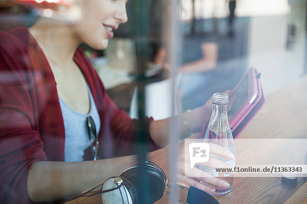 Junge Frau sitzt im Café  hält eine Flasche Wasser in der Hand und benutzt ein digitales Tablett