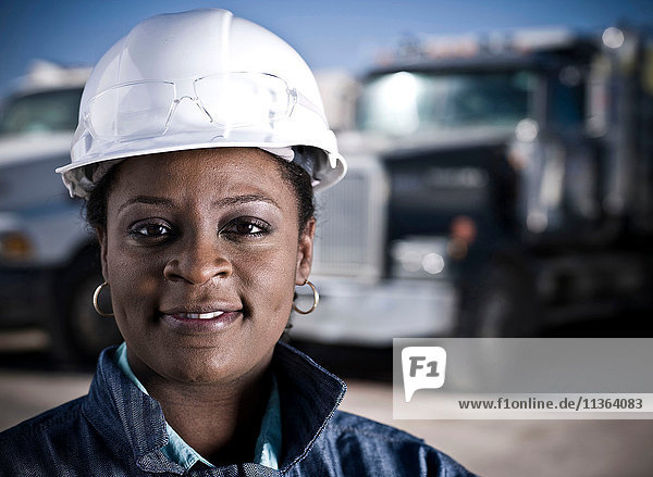 Porträt einer Arbeitnehmerin mit Schutzhelm vor den Lastwagen der Recyclinganlage