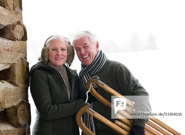 Portrait of couple standing outside cabin in snow carrying toboggan  Colorado  USA