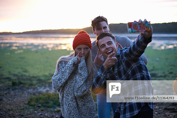 Drei junge Erwachsene nehmen Smartphone-Selfie bei Sonnenuntergang am Meer