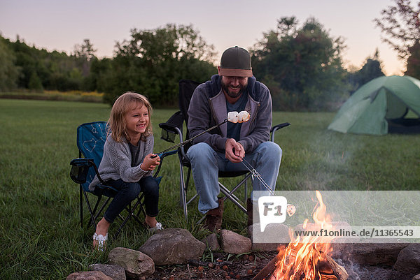 Vater und Tochter sitzen am Lagerfeuer und rösten Marshmallows über dem Feuer