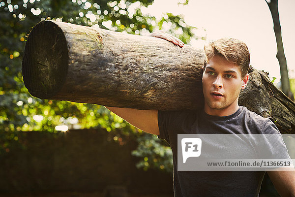 Porträt eines jungen Mannes  der im Park den Baumstamm auf die Schulter hebt.