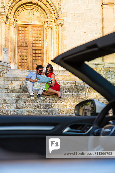 Junges Paar mit Cabriolet sitzt auf der Kirchentreppe und schaut auf die Karte  Calvia  Mallorca  Spanien