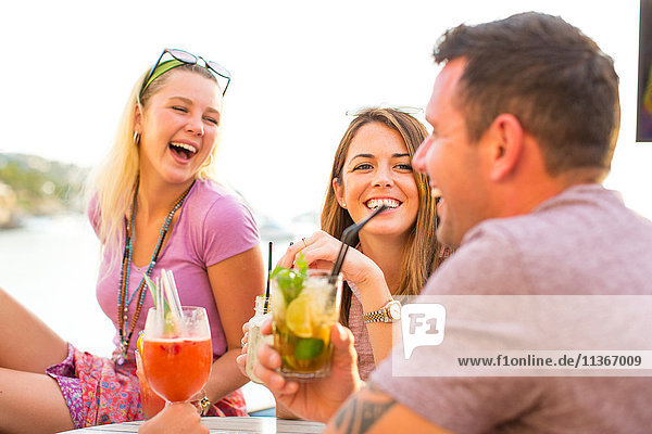 Three adult friends laughing over cocktails at waterfront restaurant  Majorca  Spain