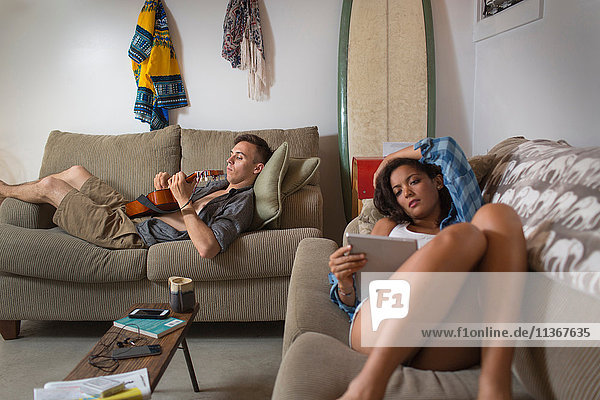 Junges Paar entspannt sich auf dem Sofa mit digitalem Tablett und spielt Gitarre