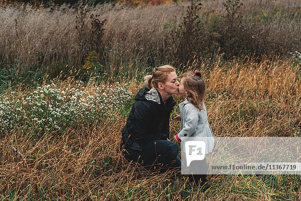 Mittelgroße erwachsene Frau küsst Kleinkind-Tochter im Feld von langem Gras