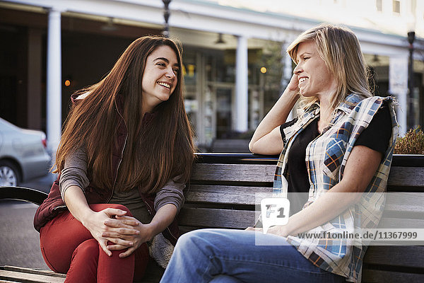 Two women sitting on a bench in an urban environment talking.