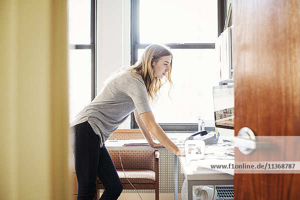 Eine junge Frau in einem Büro  die über einem Schreibtisch steht und an einem Computer arbeitet.