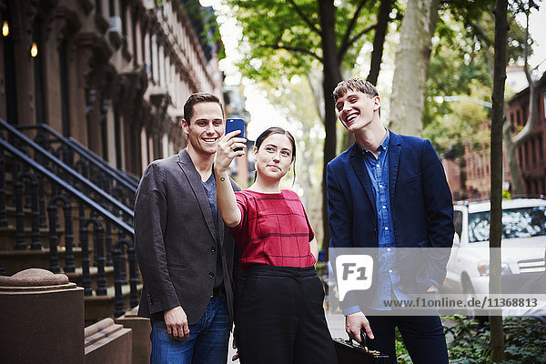 Zwei junge Männer und eine junge Frau posieren für einen Selfie  der mit einem Handy in einer Straße der Stadt aufgenommen wurde.