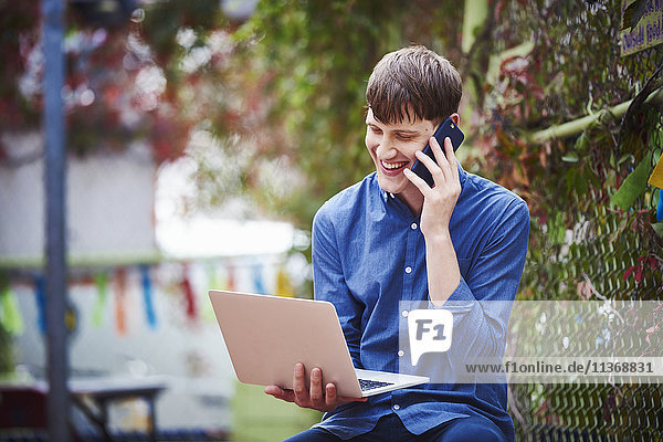 Ein junger Mann sitzt im Freien und hält einen offenen Laptop in der Hand und ein Mobiltelefon am Ohr.