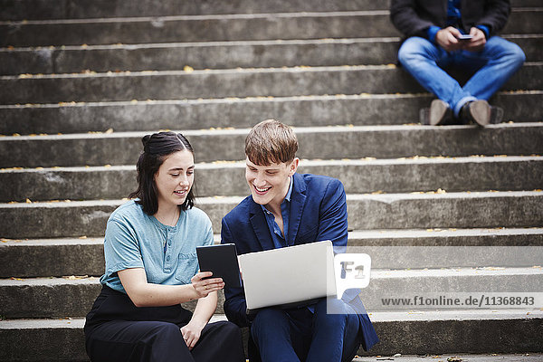 Eine junge Frau und ein junger Mann sitzen auf einer Freitreppe im Freien und schauen gemeinsam auf einen Laptop.