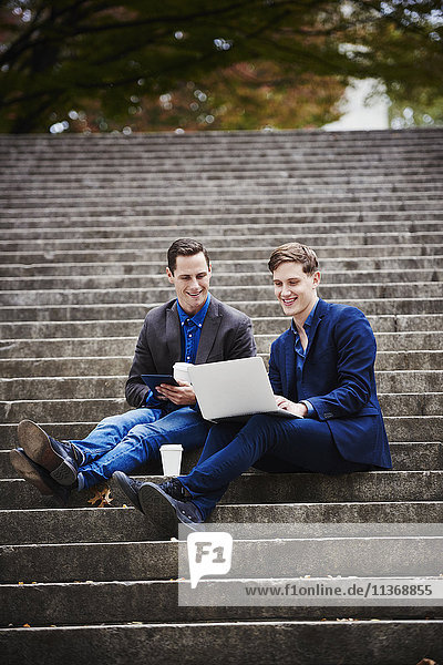 Zwei junge Männer sitzen auf einer Freitreppe im Freien und schauen gemeinsam auf einen Laptop.