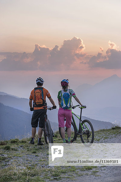 Rear view of young couple of mountain bikers standing in the alpine landscape and looking at view during sunset  Zillertal  Tyrol  Austria