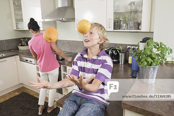 Junge jongliert mit Orangen  während die Mutter das Essen zubereitet