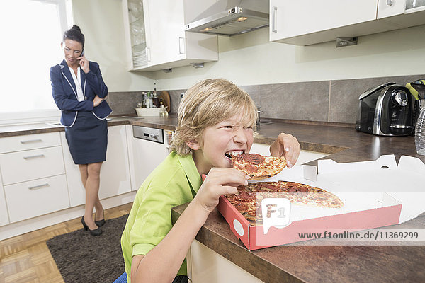 Boy eating pizza while mother has business call in kitchen