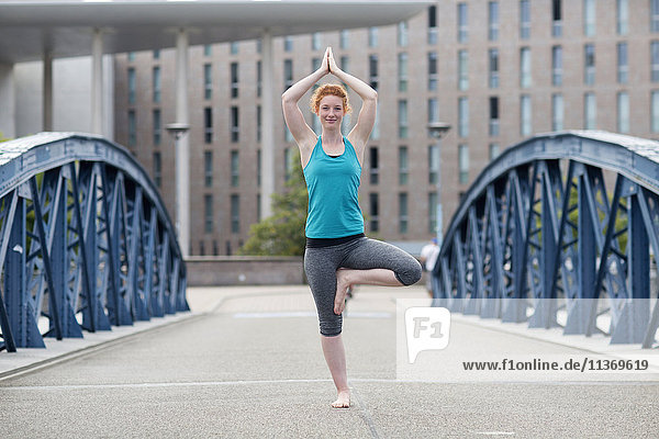 Junge Frau  die auf einer Brücke in einer Großstadt Yoga in Baumstellung macht