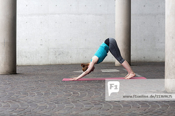 Young woman doing downward facing dog pose on exercise mat in urban city