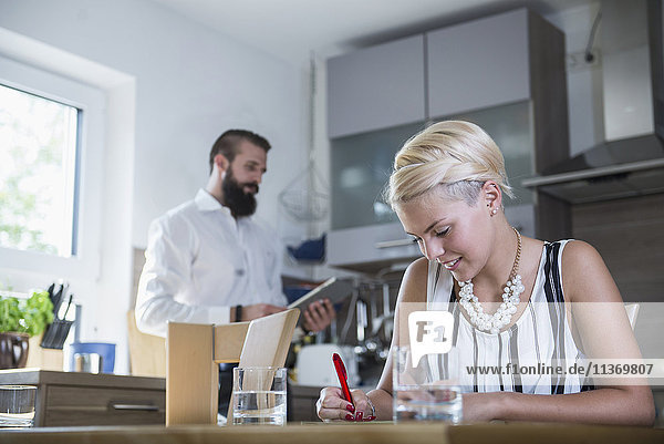 Junge Frau schreibt am Küchentisch und Mann im Hintergrund mit digitalem Tablet