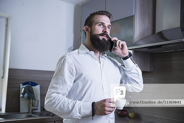 Junger Mann spricht mit seinem Smartphone und trinkt Kaffee in der Küche