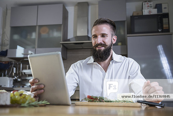 Junger Mann,  der in der Küche auf einem digitalen Tablet ein Rezept für eine Mahlzeit sucht