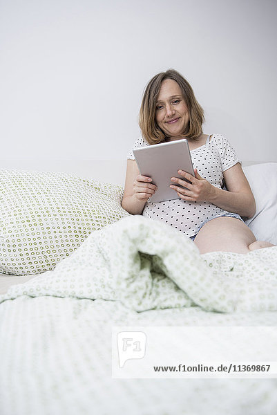 Glückliche schwangere Frau mit digitalem Tablet im Bett