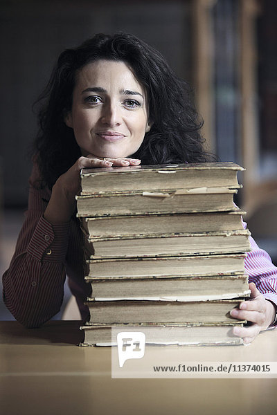 Braunhaarige junge Frau in einer Bibliothek,  deren Kopf und eine Hand auf einem Stapel alter Bücher ruhen.