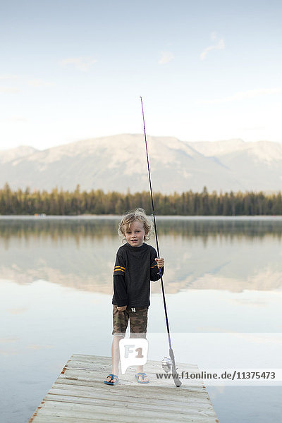 Kaukasischer Junge steht auf einem Steg am See und hält eine Angelrute