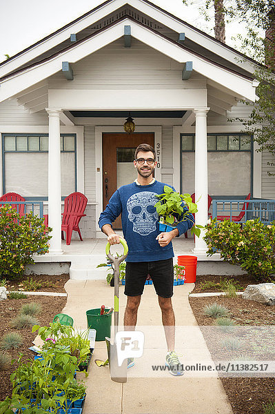 Porträt eines lächelnden hispanischen Mannes  der eine Schaufel und Pflanzen in der Nähe eines Hauses hält