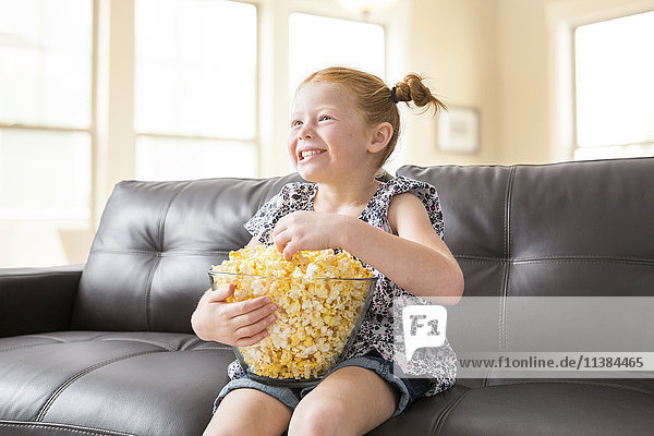 Kaukasisches Mädchen sitzt auf dem Sofa und isst eine Schüssel Popcorn