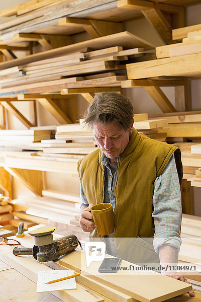 Kaukasischer Tischler trinkt Kaffee und benutzt sein Handy in der Werkstatt